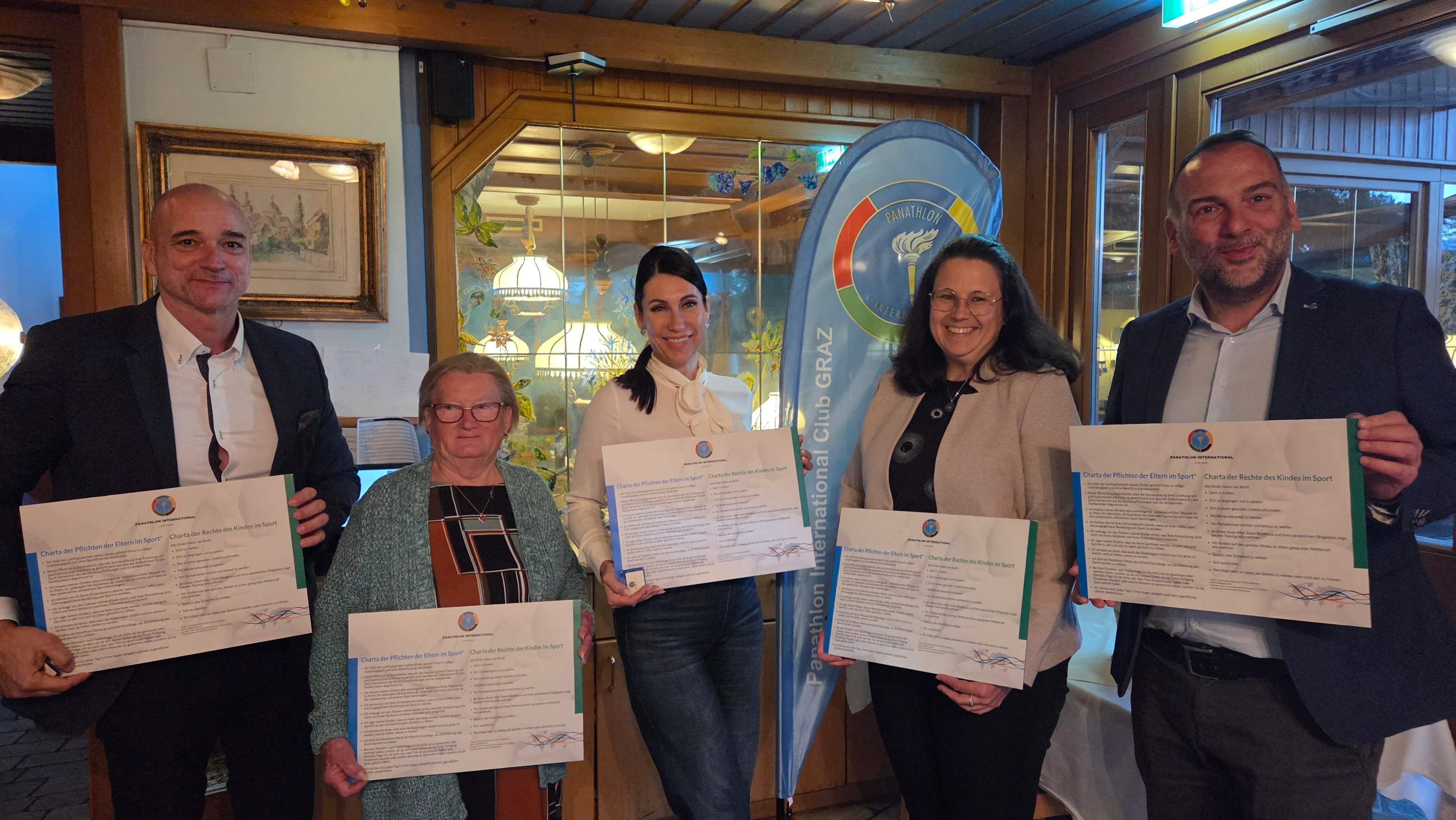 Fünf neue Mitglieder des Panathlon Club Graz halten die Urkunden der Charta der Rechte und Pflichten im Sport, im Hintergrund die Panathlon-Beachflag im Restaurant Häuserl im Wald